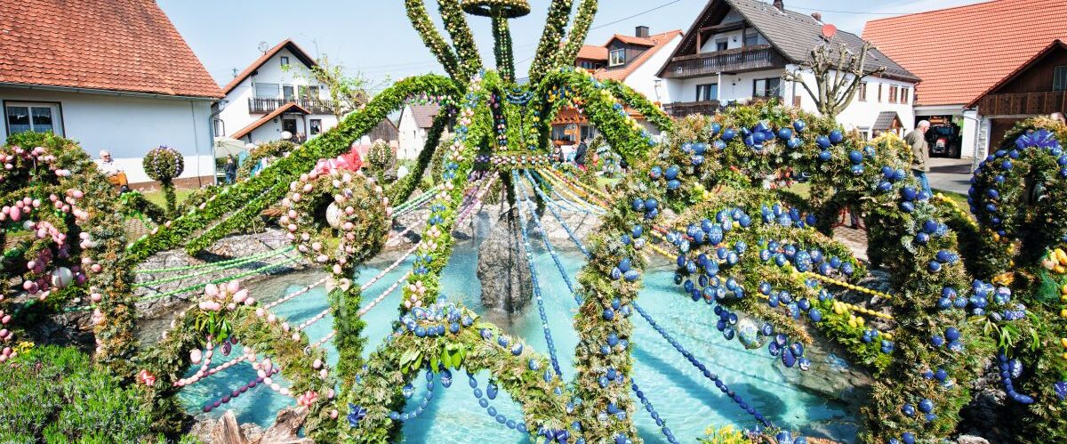 Ein Dorfbrunnen ist reich geschmückt mit grünem Tannengrün, bunten Ostereiern und Girlanden.